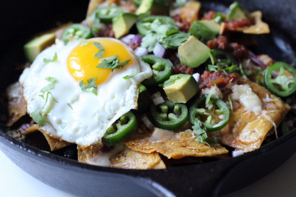 Close up of tortilla chips in a cast iron pan, with jalapenos, salsa, avocado, cilantro, and a fried egg on top.